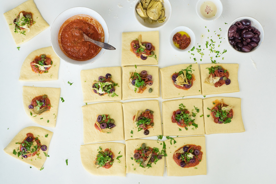 12. Die Teig-Quadrate mittig mit einem Teelöffel Tomatensauce sowie getrockneten Tomaten, Oliven, Artischocken, Petersilie und Kapern belegen.